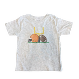 A light heather gray baby t-shirt laid flat on a dark patterned rug. The shirt features a graphic of a yellow chair with a brown football and an orange duck wearing a football helmet.