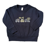 A navy blue long-sleeve sweatshirt for toddlers, featuring a graphic of three construction vehicles (a dump truck, a crane, and a cement mixer) on the chest. The sweatshirt is laid flat on a blue and beige patterned rug.