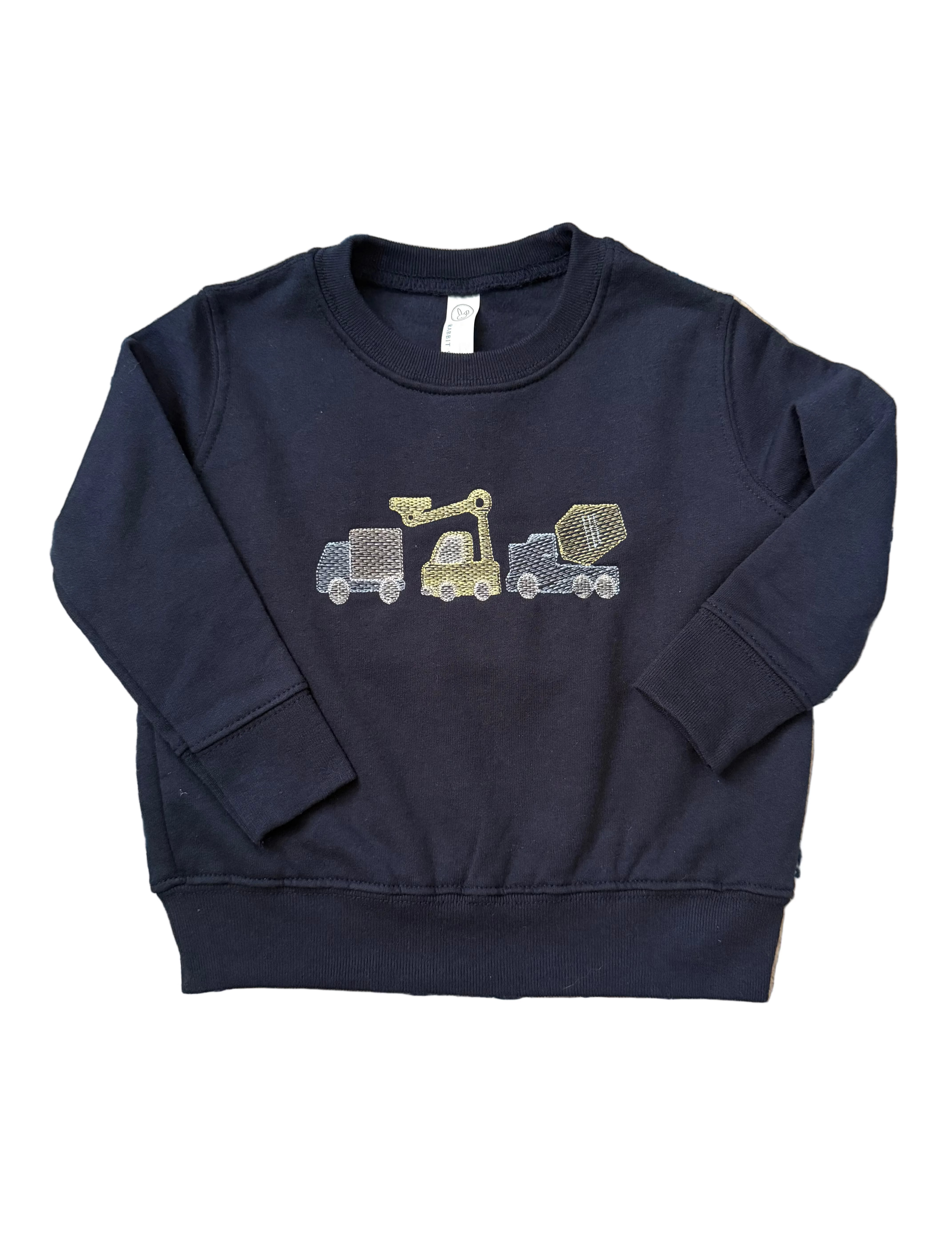 A navy blue long-sleeve sweatshirt for toddlers, featuring a graphic of three construction vehicles (a dump truck, a crane, and a cement mixer) on the chest. The sweatshirt is laid flat on a blue and beige patterned rug.