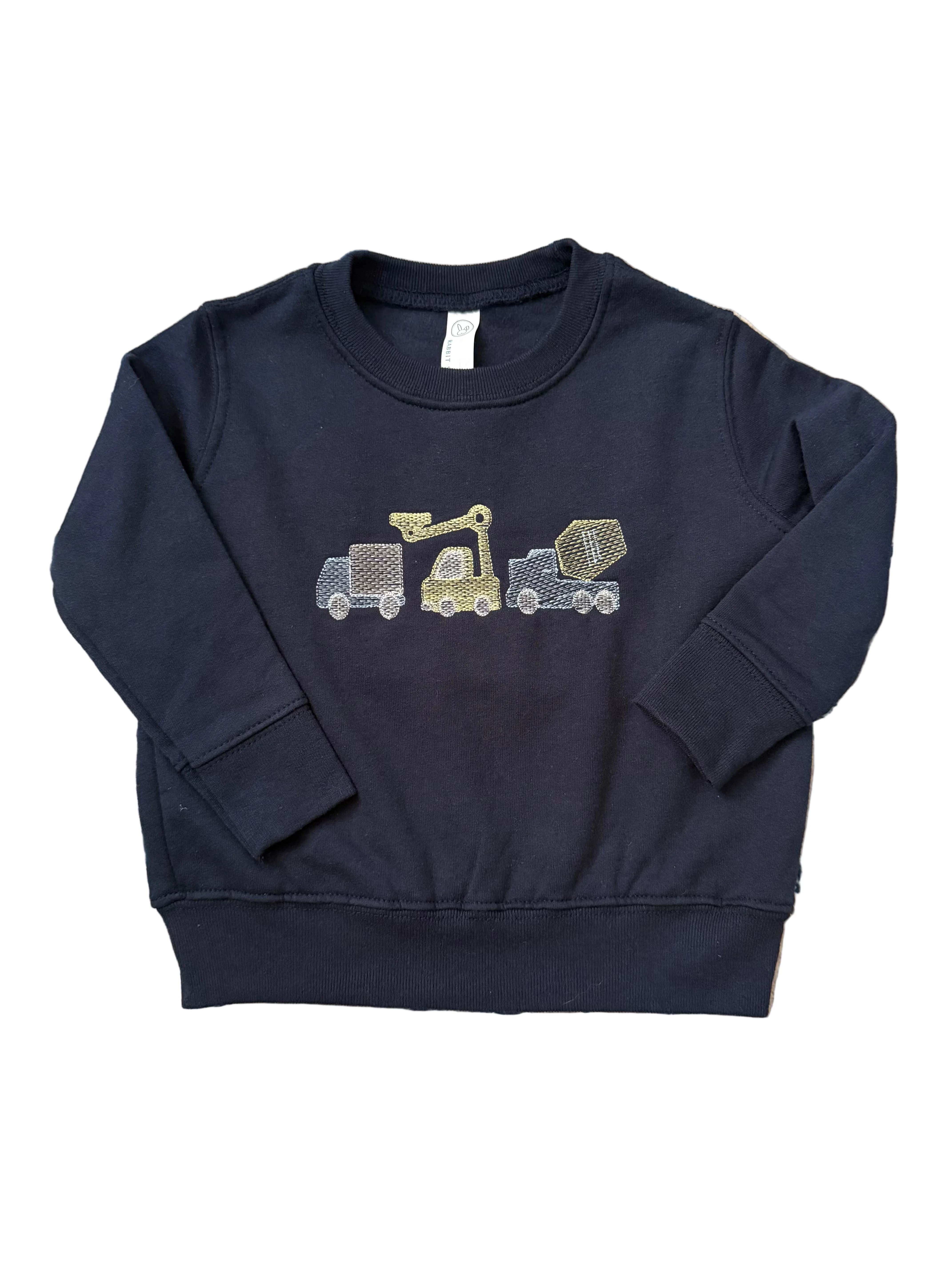 A navy blue long-sleeve sweatshirt for toddlers, featuring a graphic of three construction vehicles (a dump truck, a crane, and a cement mixer) on the chest. The sweatshirt is laid flat on a blue and beige patterned rug.