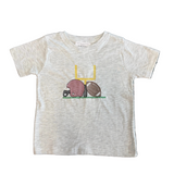 A toddler's short-sleeve heather gray t-shirt featuring a football-themed graphic with a pink helmet, brown football, and yellow goalpost, laid flat on a patterned gray rug.