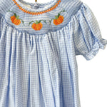 A light blue and white gingham baby dress with orange and green pumpkin embroidery and a smocked neckline, hanging on a gold hanger against a white lattice screen.