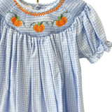 A light blue and white gingham baby dress with orange and green pumpkin embroidery and a smocked neckline, hanging on a gold hanger against a white lattice screen.