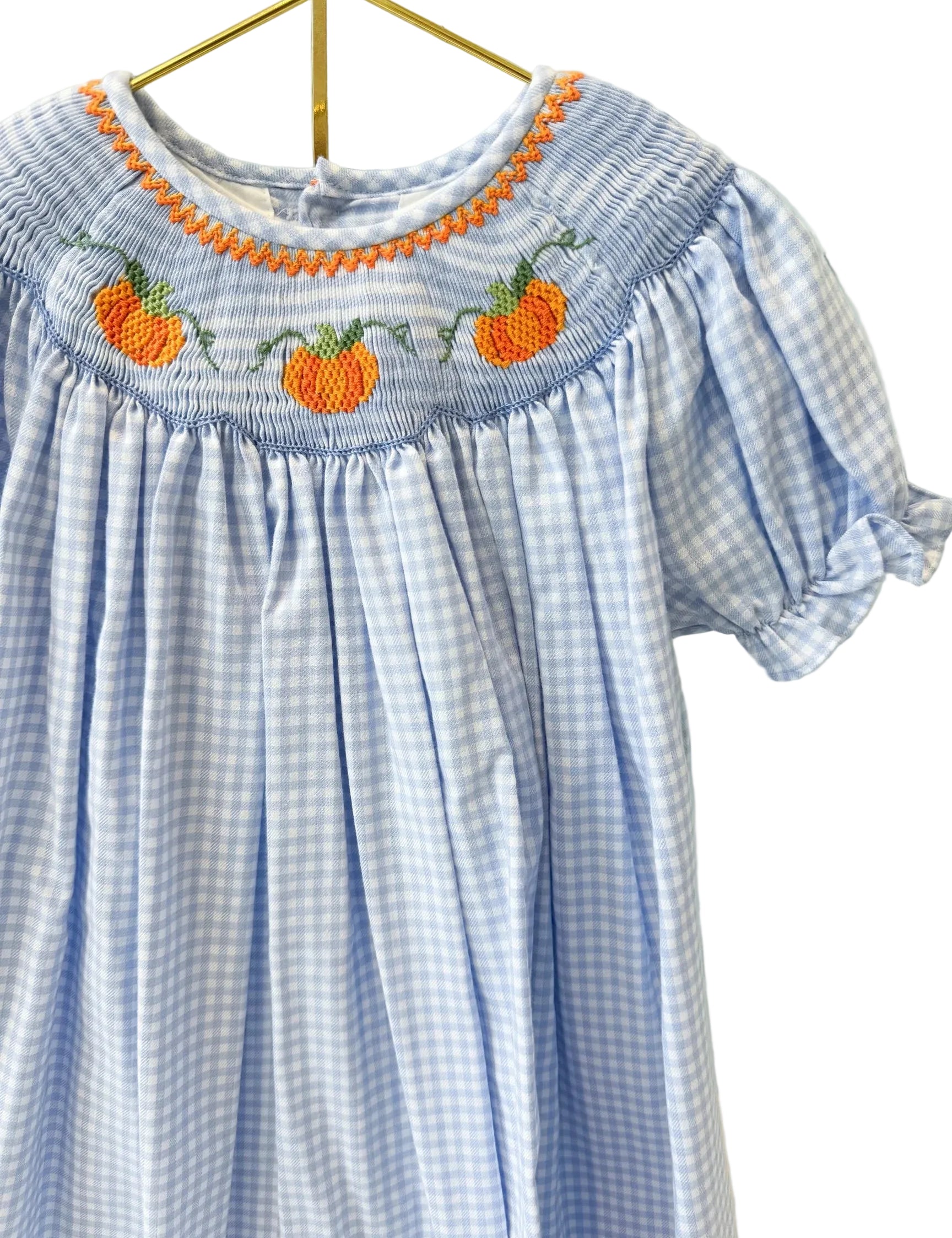 A light blue and white gingham baby dress with orange and green pumpkin embroidery and a smocked neckline, hanging on a gold hanger against a white lattice screen.