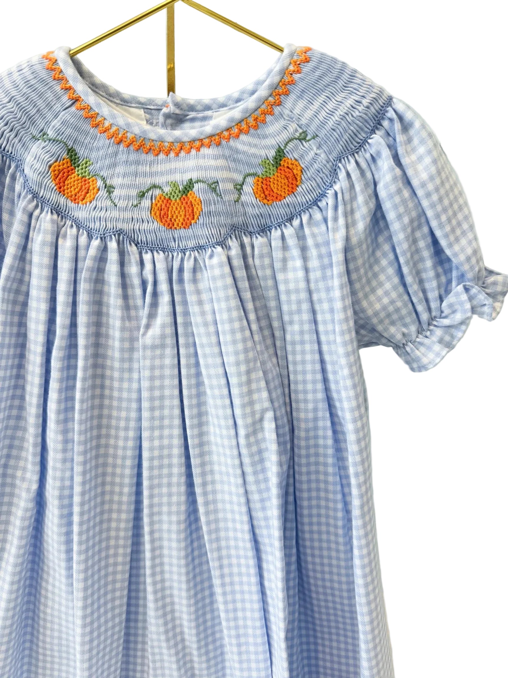 A light blue and white gingham baby dress with orange and green pumpkin embroidery and a smocked neckline, hanging on a gold hanger against a white lattice screen.